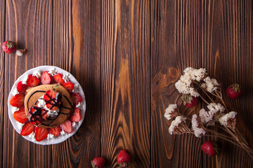 Homemade pancakes with strawberries, whipped cream and chocolate topping, decorated with flowers on a brown wooden background. Overhead shot. Flat lay. Copy space
