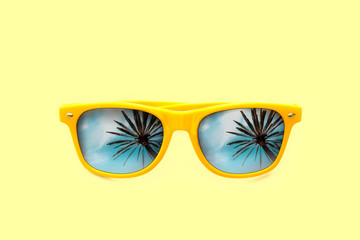 Summer concept image: yellow sunglasses with palm tree reflections isolated in pastel yellow background. Minimalist image ready for summer, sun protection, hot days and tropical travel vacation.