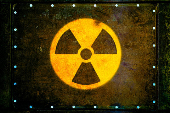 Round yellow radioactive (ionizing radiation) danger warning symbol painted on massive rusty metal plate fixed with metallic rivets to the wall with dark rusty brown and green moss.