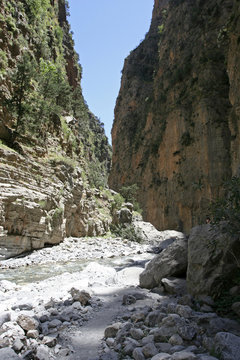 The gorge with high steep rocky slopes with green plants, the small  mountain river and the white stony path on its bottom on the sunny day.