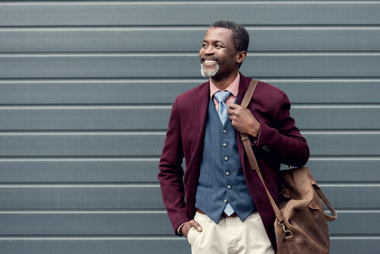 happy stylish african american man in burgundy jacket with leather bag