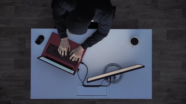 Young man in black hoodie stretching fingers and typing on laptop and looking at monitor during night, sitting behind white desk, top shot