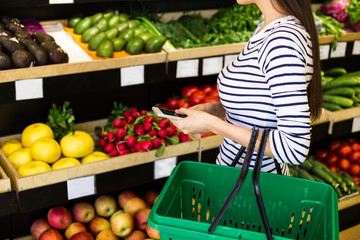 Attractive smiling young girl in casual clothes selects fruits and vegetables from a shelf in a grocery store.