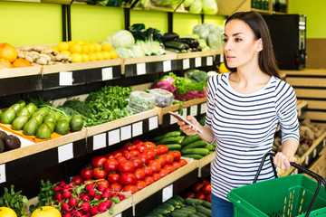 Attractive smiling young girl in casual clothes selects fruits and vegetables from a shelf in a grocery store.