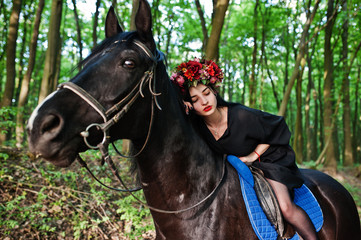 Mystical girl in wreath wear in black at horse in wood.