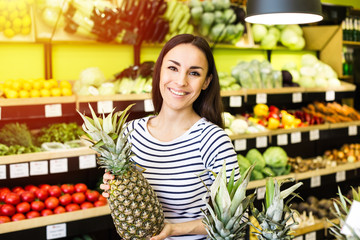 Attractive smiling young girl in casual clothes selects fruits and vegetables from a shelf in a grocery store.