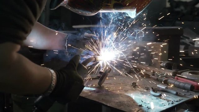 Close up of woman welder or blacksmith wearing protection mask repairing and welding metal object that makes shower of sparkles in her workshop