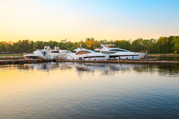 Fototapeta premium Luxury yachts docked in marina at colorful sunset. Marine parking, modern motor boats moored in water bay at pier. Summer view of river bank, embankment and superyachts in sunshine. Sunny evening.