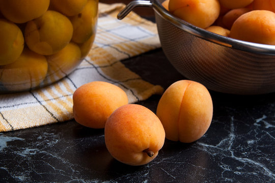 Fresh organic harvested apricots in steel colander and big glass jar with canned apricot compote on dark marble..