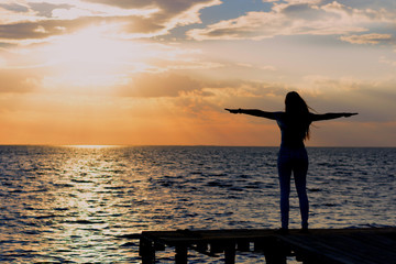 Fototapeta premium A young slender girl stands on a wooden bridge (fishing bridge) spreading her hands over the sea and looking at the sunset (dawn) of the sun. Life and freedom. 