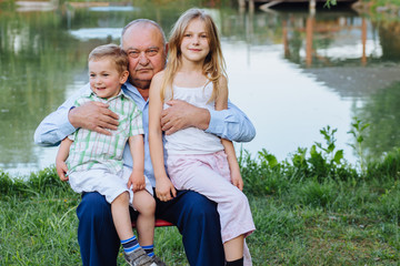 Fototapeta premium Sweet family portrait of three happy relatives - grandfather hugging with his granddaughter and grandson over lake background at summer time.