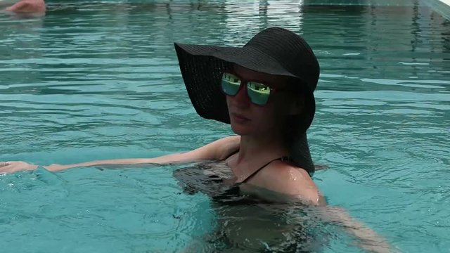 young woman in a black broad-brim and sunglasses resting in swimming pool