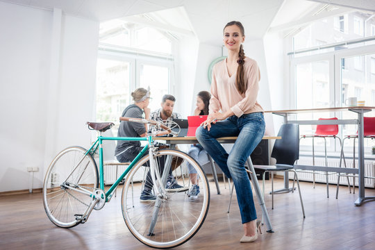 Full length of a beautiful and confident young woman wearing blue jeans, while sitting on a desk near her commuter bike in a co-working office space