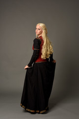 Fototapeta premium full length portrait of pretty blonde lady wearing a red and black fantasy medieval gown. standing pose on grey background.