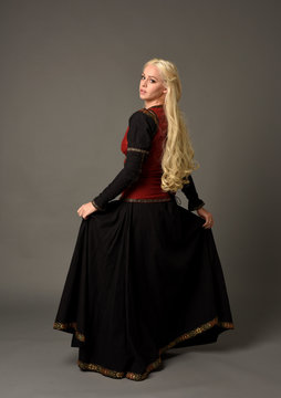 full length portrait of pretty blonde lady wearing  a red and black fantasy medieval gown. standing pose on grey background.