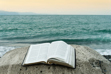 Bible open on top of a large stone in front of the green sea and an orange sky. Copy space.