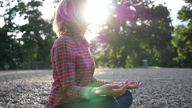 Young woman meditating relaxing sitting in a park on glade pebbels ground in sunset sun rays