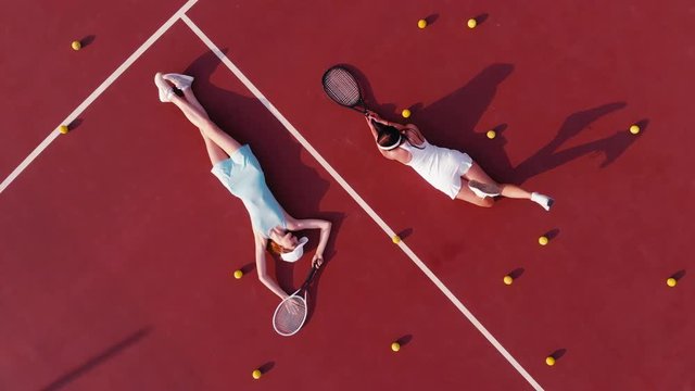 OVERHEAD DRONE shot of two young Caucasian teen models wearing fashionable tennis dresses, lying on tennis hardcourt with a lot of balls, summer sunny day outdoors. Fashion portrait shoot