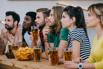 group of multicultural friends drinking beer and watching soccer match at bar