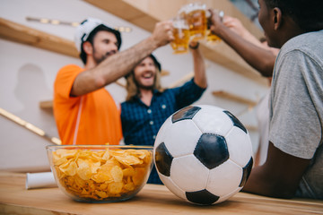 closeup view of soccer ball and bowl with chips with group of football fans celebrating and clinking by beer glasses behind at bar