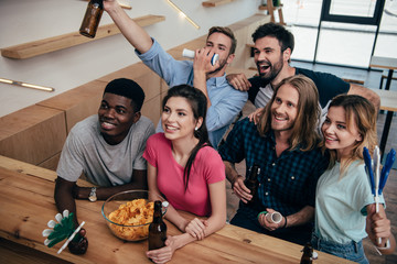 high angle view of excited multicultural friends celebrating with fan horns and hand clappers during watch of soccer match at bar