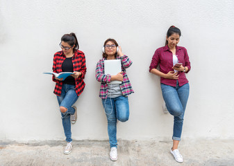 Group of friend standing and laying against the white wall outdoor, reading a book, checking news from cell phone, and holding a laptop and listening to music.