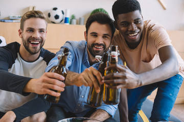 happy young multicultural friends clinking beer bottles and celebrating during watch of soccer match at home
