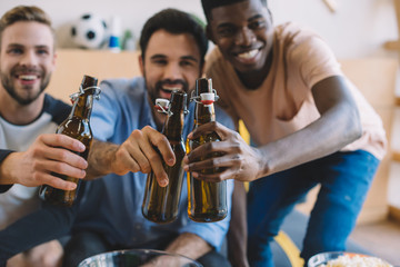 excited young multicultural friends clinking beer bottles and celebrating during watch of soccer match at home