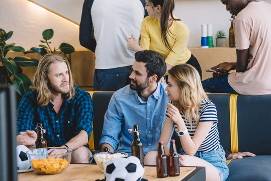 cropped image of group multicultural football fans sitting on sofa and talking to each other near table with beer, chips and popcorn