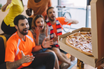 close up shot of pizza in box and excited group of friend sitting on sofa at home