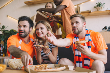 young man pointing by hand and his friends eating pizza and watching soccer match at home