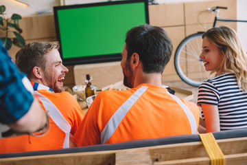 back view of smiling friends in orange fan t-shirts celebrating and watching football match on tv screen at home