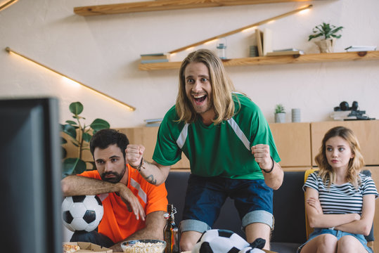 Happy Young Man In Green Fan T-shirt Celebrating While His Upset Friends Sitting Behind On Sofa During Watch Of Soccer Match At Home