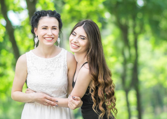 Portrait of two young beautiful women laughing and hugging each other in green summer park. Pretty females bride and bridesmaid smiling and looking into camera. Copy spece
