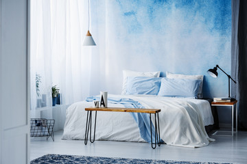 Lamp on wooden table next to a blue and white bed in bedroom interior with ombre wall