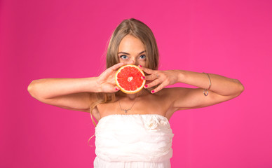 Beautiful blonde woman with a grapefruit in hands on a pink background. Female with one half of tasty juicy grapefruit closes his mouth to them.