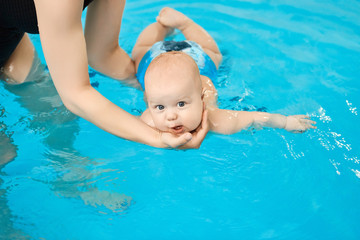 Little baby swimming in water pool with help from mothers hands.