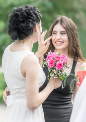 Portrait of two young beautiful women friends. Pretty bride with roses bouquet helping her smiling bridesmaid. Wedding at Sunny summer day in green park