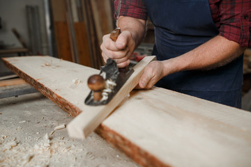 Master carpenter in shirt and apron strokes  plane in workshop. Close up