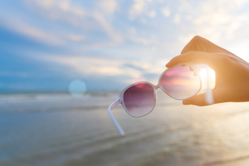 Fototapeta na wymiar Soft focus on woman hand holding sunglasses over sea and Sandy beach in background during sunset for summer holiday and vacation concept.