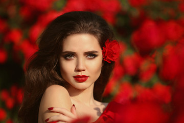 Naklejka premium Outdoors portrait of Natural Beauty woman in red roses. Sensual female with rose flowers in hair. Sexy brunette with makeup over sunset blossom park. Passion and expression. Spanish flamenco style.