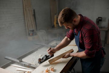 Master carpenter in shirt and apron strokes  plane in workshop. Close up