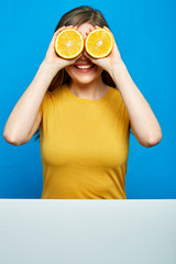 Smiling woman holding two slices orange in front of eyes.