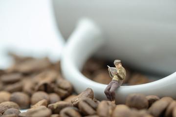 Business, reading and relaxation concept. Businessman miniature figures sitting and reading a newspaper or book on handing of mug cup of coffee with coffee beans.