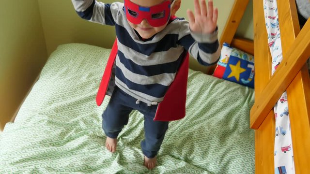 A cute little boy with a super hero cape and mask jumping on the bed