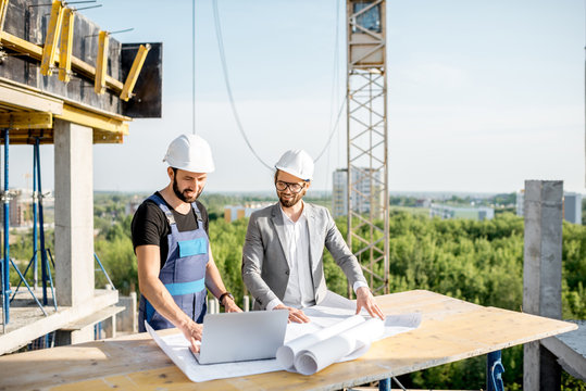 Engineer with worker in uniform working with architectural drawings and laptop at the table on the construction site outdoors