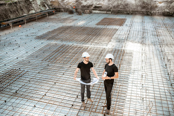 Two builders in t-shirts and protective helmets standing with drawings on the concrete foundation at the construction site