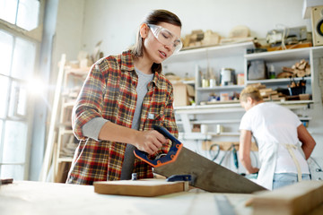 Obraz premium Adult woman in safety goggles using saw to cut timber board while working in carpenter workshop
