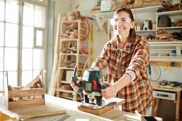 Pretty female carpenter looking at camera and using electric saw to cut wooden plank while working in workshop. 