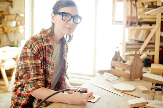 Adult woman in glasses using nice pyrography burner and looking at camera while sitting at workbench in carpenter workshop. 